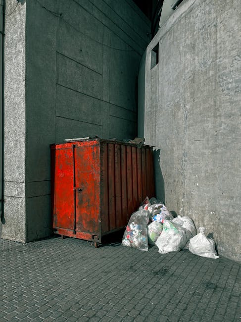 A large, industrial red metal waste skip with a rusted, weathered finish is positioned against a dark green wall and a light grey concrete wall in an outdoor urban setting. The skip's surface shows si