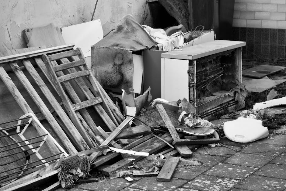 A pile of mixed rubbish, including cardboard boxes, paper packaging, and plastic bags, is gathered against a weathered brick and concrete wall outdoors. The cardboard boxes are various sizes, some par